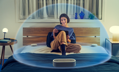 Image d'une personne assise sur un lit, entourée du système HT-AX7 avec une bulle transparente représentant le son spatial qui enveloppe la personne