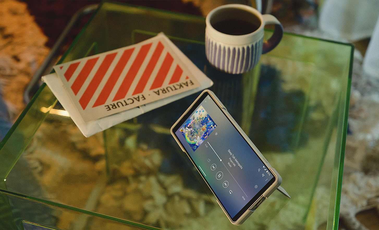 Image of an Xperia 5 V being propped up by its stand on a glass coffee table with a music playing interface on screen