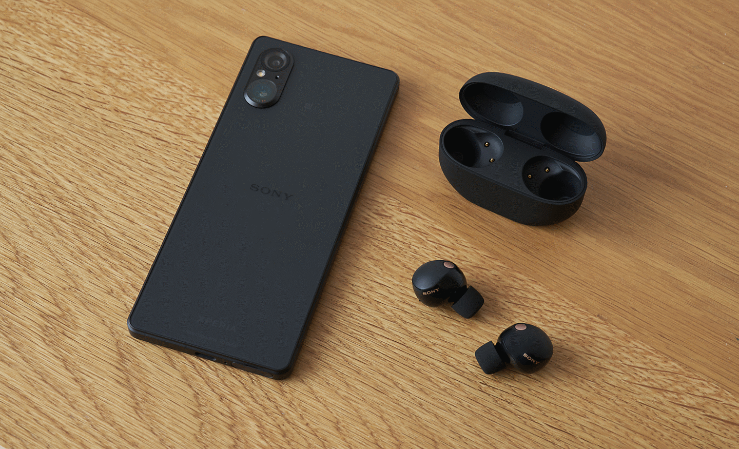 A black Xperia 5 V face down on a wooden table next to a pair of black in-ear headphones and their open case