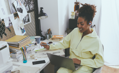 Image of a woman sitting at a busy desk using a laptop and wearing lavender WF-C700N Wireless Noise Cancelling headphones