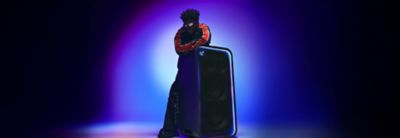 Photograph of a person with short spiky dark hair and sunglasses, wearing a black jacket with red sleeve stripes. The person leans on a ULT TOWER MAX speaker with illuminated blue and purple LED trim and a circular ULT logo, against a pastel gradient background.