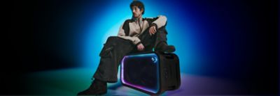 Photograph of a person with short curly dark hair sitting on a ULT TOWER 7 speaker. The horizontal speaker features illuminated blue and pink LED trim around its front grille and a circular ULT logo. The person wears a two-tone jacket and dark trousers against a blue gradient background.