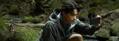 Photo of a person in a grey waterproof jacket crouching in a wooded area, holding a smartphone with an ECM-AX10 microphone to record.