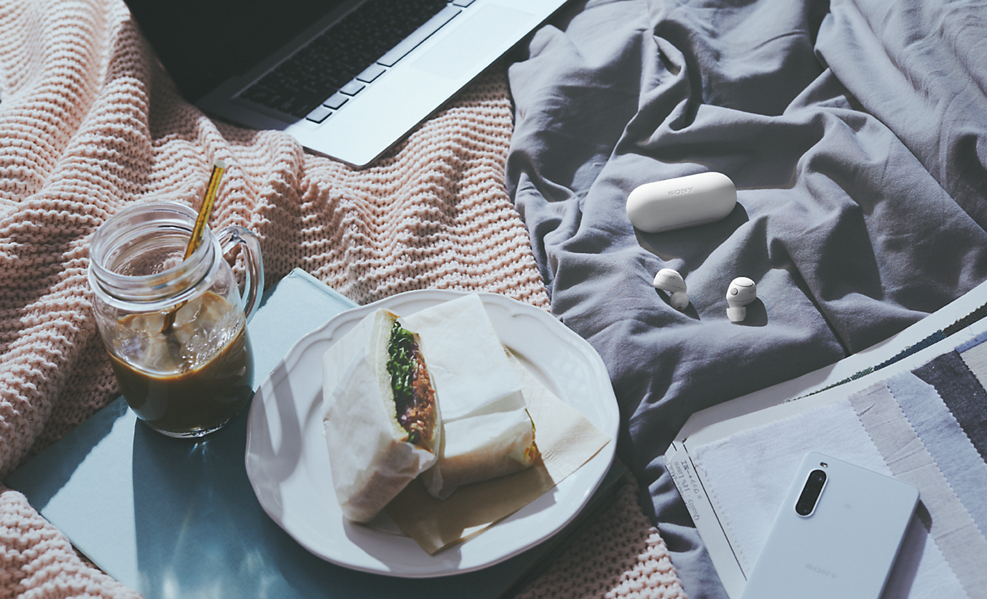 Image of the white WF-C700N headphones and case on a picnic blanket with food, drink, a laptop and a phone