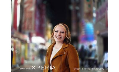 Человек, четко сфокусированный на камере и улыбающийся на фоне размытого ночного города. Фотография сделана на Xperia.