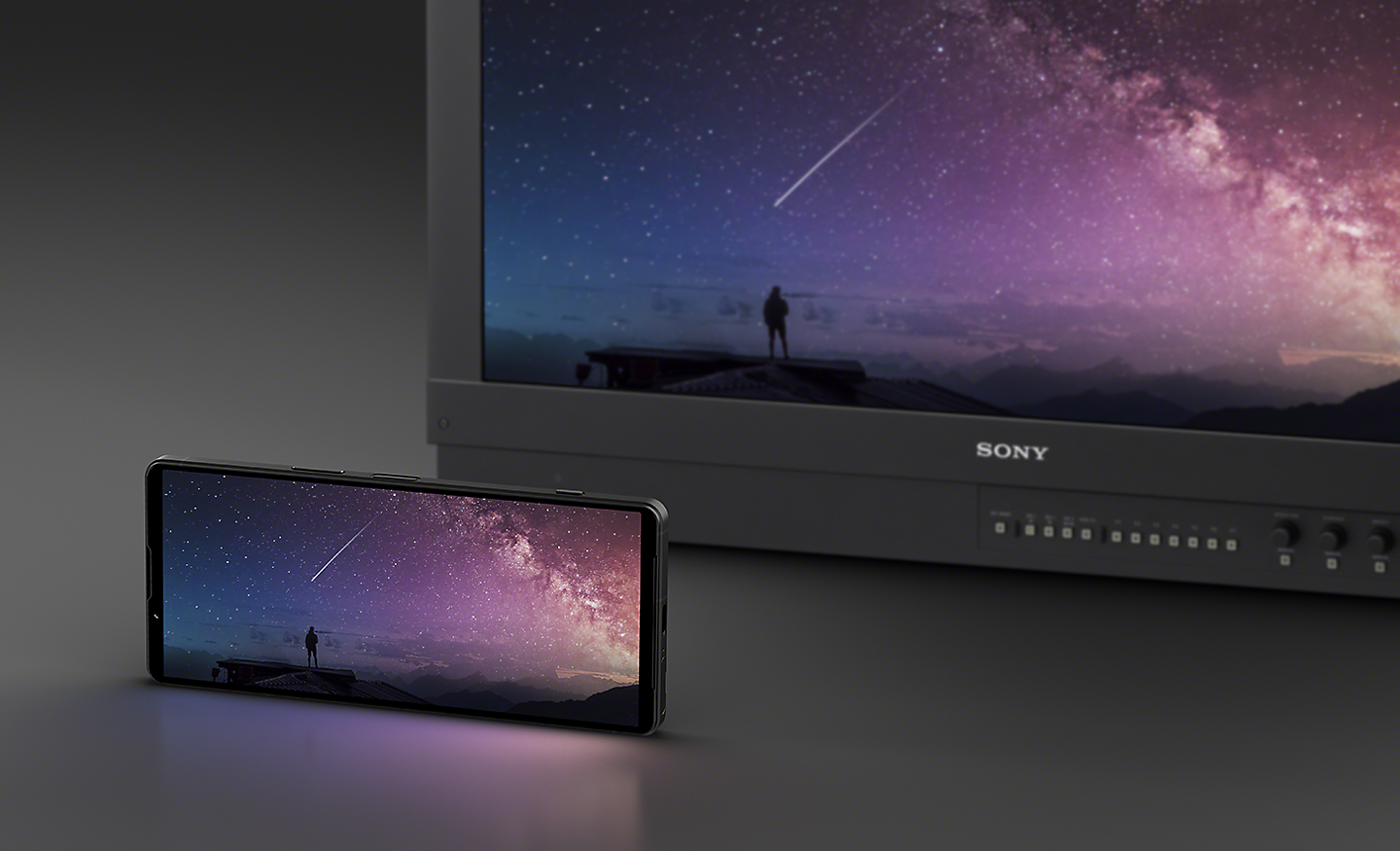 Xperia 1 V ในตำแหน่งแนวนอนทางด้านหน้าจอภาพสีเพื่อมืออาชีพของ Sony โดยทั้งสองสิ่งแสดงภาพท้องฟ้ายามค่ำคืนเหมือนกัน