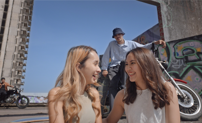 An urban setting, two young people in the foreground, two young people on motorbikes in the background, and bright blue sky