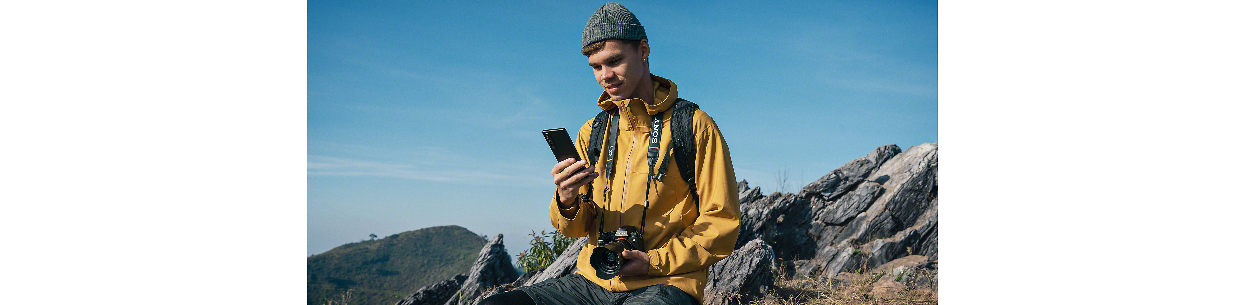 En person i ett bergigt landskap – han har en Sony Alpha-kamera runt halsen och tittar på sin Xperia 1 V