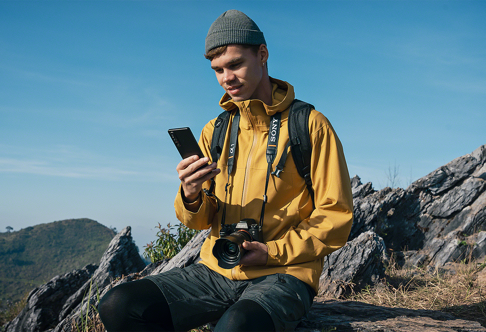 En person i ett bergigt landskap – han har en Sony Alpha-kamera runt halsen och tittar på sin Xperia 1 V
