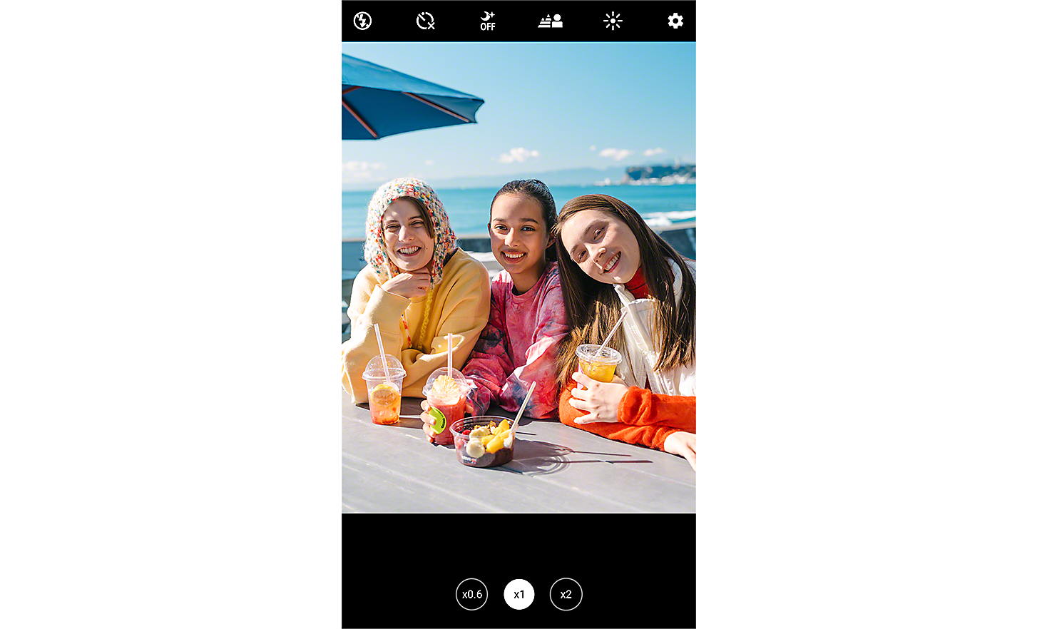 Skjermbildet viser et bilde av tre unge kvinner som sitter ved et bord med utsikt over havet og smiler til kamera