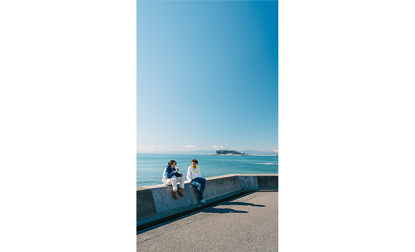 Dvaja ľudia sedia na nízkej stene pred širokým pohľadom na oceán