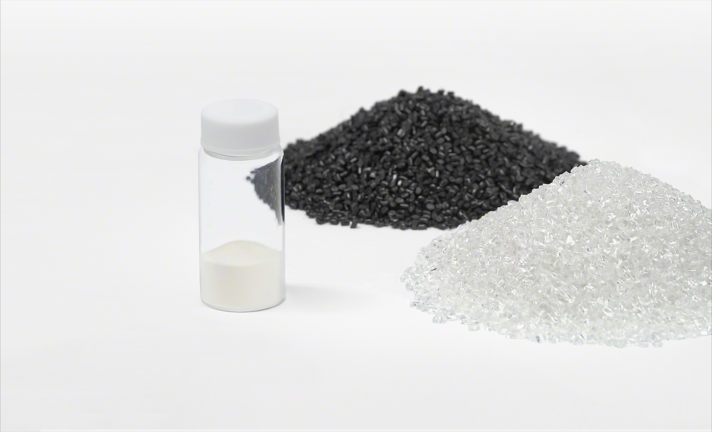 A vial of recycled plastic material alongside a pile of black plastic fragments and a pile of white plastic fragments