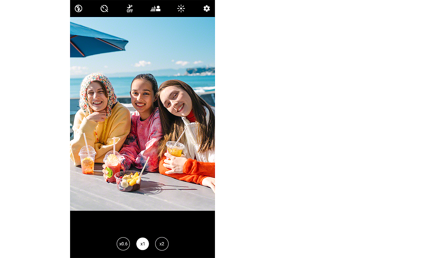 Snímka obrazovky, ktorá zobrazuje obrázok troch mladých žien, ako sa usmievajú do fotoaparátu, sedia pri stole s výhľadom na oceán