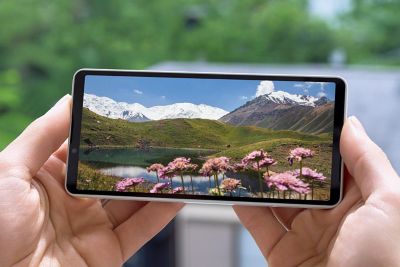 Dos manos sostienen un smartphone en horizontal, mirando una foto panorámica de montañas, un lago y flores rosas en la pantalla, visible bajo la intensa luz solar.