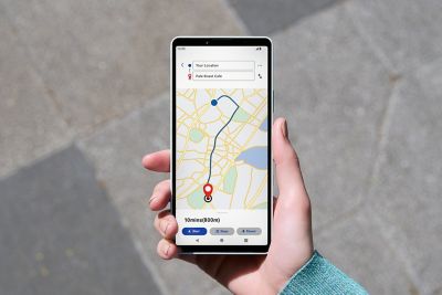 Una mano sostiene un smartphone mostrando un mapa de navegación con una ruta que va desde un marcador de inicio azul hasta un marcador de destino rojo, visible bajo la intensa luz solar.