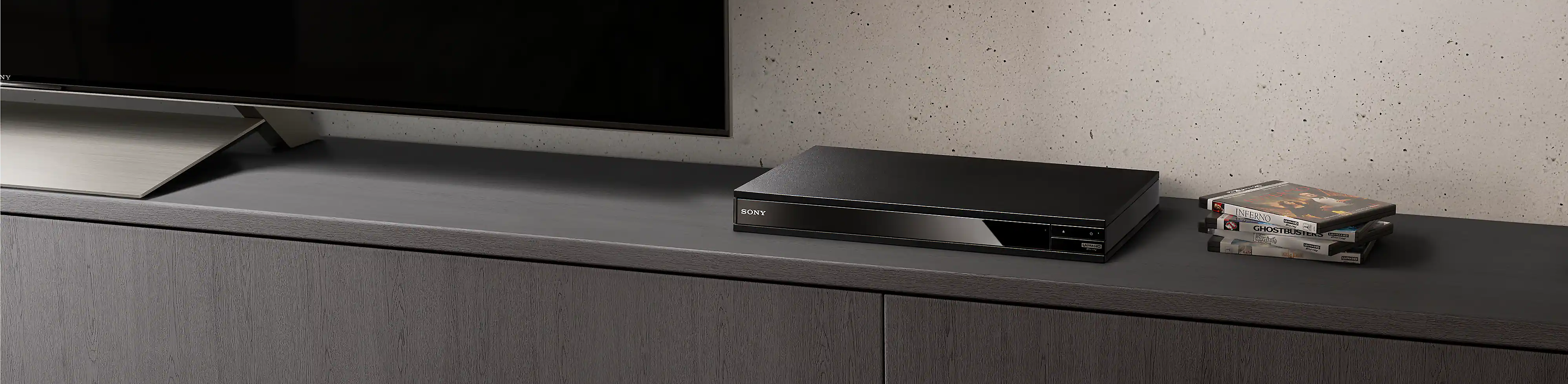 Ein schwarzer Blu-ray Disc Player zwischen einem Stapel von Blu-ray Discs und einem schwarzen Fernseher.