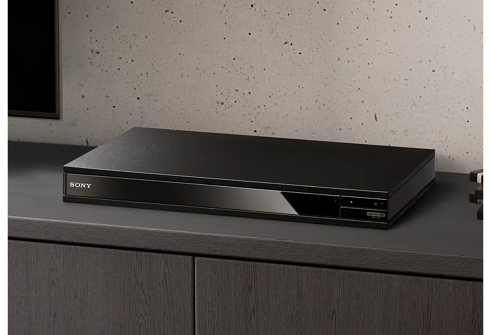 Црн Blu-ray Disc-плеер се гледа помеѓу куп Blu-ray-дискови и црн телевизор.