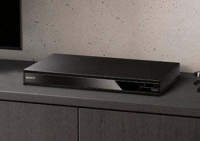 A black Blu-ray disc player is seen between a stack of blu-ray discs and a black TV.