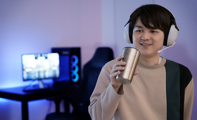 Imagen de un hombre bebiendo de una taza de metal mientras usa unos audífonos con micrófono para juegos INZONE H9 con una PC de fondo