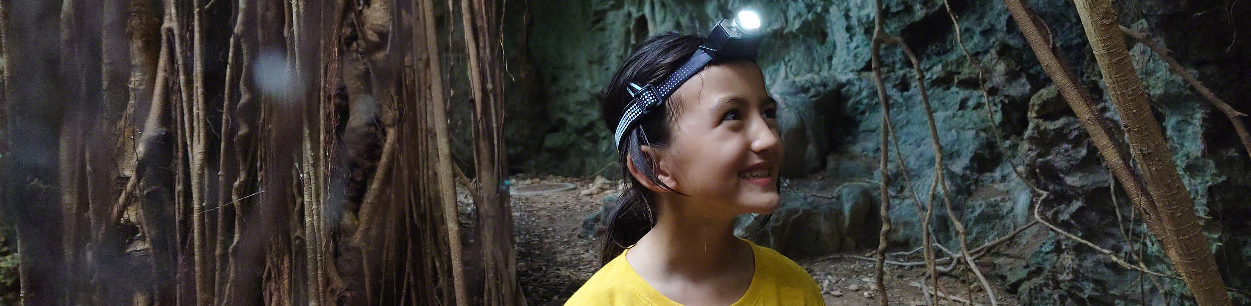 Dievča v žltom tričku s baterkou na hlave sa prechádza medzi stromami a skalami