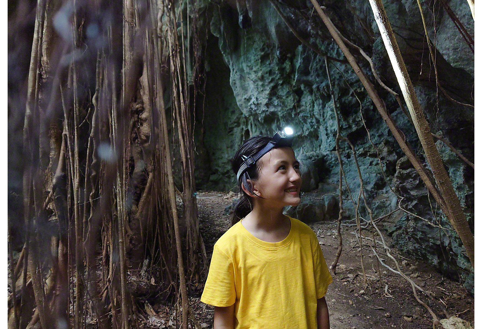 女孩穿著黃色 T 恤，頭上戴著電筒，在樹木之間和岩石表面行走