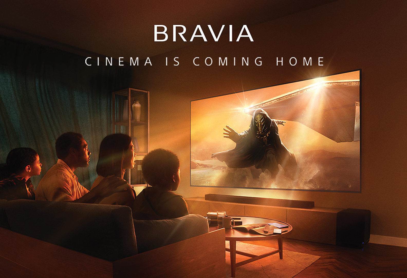 Living room scene with four people sitting on a sofa and wall-mounted TV and soundbar with screenshot of a futuristic masked and cloaked villain stepping through orange mist with spaceship in the background, to the bottom of the image the words BRAVIA and cinema is coming home