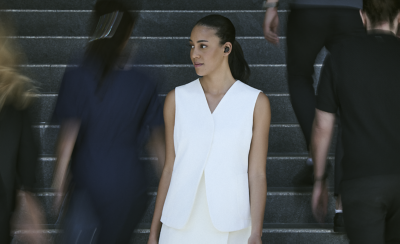 흰색 조끼와 치마를 입고 왼쪽 귀에 블랙 이어버드를 착용한 젊은 여성이 계단을 내려가고, 그녀를 지나 계단을 올라가는 사람들의 모습은 흐리게 처리되어 있습니다.