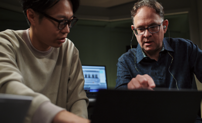 스튜디오 분위기의 장소에서 안경을 쓴 두 남성이 앉아 노트북을 가리키며 보고 있고 배경에는 화면이 추가로 보입니다.
