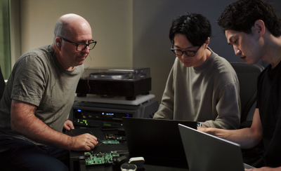 스튜디오 분위기의 장소에서 남성 세 명이 앉아 노트북을 보고 있습니다. 전경에는 프로세서와 이어버드 한 쌍이 있고 배경에는 턴테이블과 기타 오디오 장비가 있습니다.