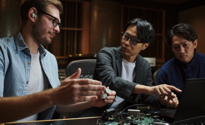 책상에 앉아 노트북을 바라보고 있는 세 명의 남성 앞에 프로세서와 이어버드가 놓여 있습니다. 한 남성은 이어버드를 착용하고 있고 다른 남성은 화면을 가리킵니다.