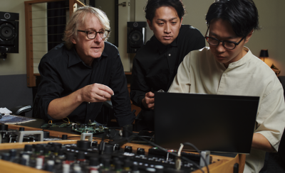 스튜디오 분위기의 장소에서 믹서 뒤로 젊은 남성 두 명과 나이 든 남성 한 명이 앉아 노트북을 보고 있습니다. 배경의 벽에는 스피커가 있습니다.