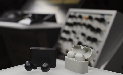 오디오 장비를 배경으로 탁자 위에 놓인 두 개의 소니 이어버드 케이스에 초점이 맞춰져 있습니다. 블랙 케이스가 닫혀 있고 테이블 위에 이어버드가 있으며 열려 있는 화이트 케이스에 이어버드가 보입니다.