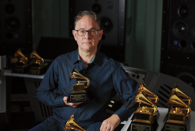 안경을 쓰고 파란색 셔츠를 입은 중년 남성이 스튜디오 분위기의 장소에서 그래미상을 들고 다른 7명에게 둘러싸여 있습니다.