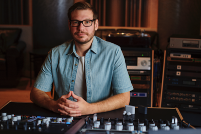 데님 셔츠와 흰색 티셔츠를 입은 남자가 스튜디오의 믹싱 테이블 앞에 손깍지를 끼고 앉아 있습니다.