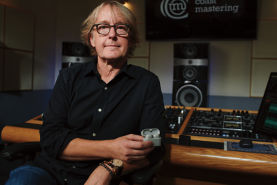 스튜디오 분위기의 장소에서 검은색 셔츠를 입은 한 남성이 의자에 앉아 화이트 이어버드가 담긴 케이스를 들고 있습니다. 남성 뒤쪽 벽에는 스피커가 있고 "coast mastering"이라고 표시되어 있습니다.