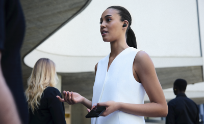 한 여성이 흰색 조끼를 입고 왼쪽 귀에 블랙 이어버드를 착용하고 있습니다. 그녀는 바깥에서 손에 전화기를 들고 있고, 주위에 사람이 여러 명 있습니다.