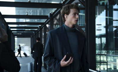 한 젊은 남성이 화이트 이어버드를 착용한 채 전화기를 들고 현대적인 유리 건물에서 걷고 있습니다. 배경에서 사람들이 여행 가방을 끌고 있습니다.