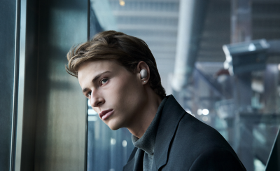 En ung mann iført svart jakke og grå genser med rullekrage ser til venstre for kameraet med en hvit ørepropp i det venstre øret.