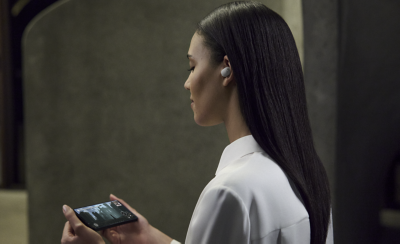 카메라 반대쪽을 바라보며 화이트 이어버드를 왼쪽 귀에 꽂은 채 휴대폰으로 동영상을 시청하는 젊은 여성.