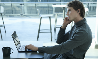 شاب يرتدي بلوزة عنق رمادية يعمل على جهاز كمبيوتر محمول في مقهى، ويرتدي سماعات أذن صغيرة وينظر إلى الأفق. إعداد بسيط.