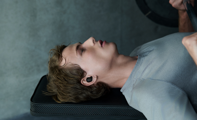 한 젊은 남성이 체육관에서 벤치에 누워 체스트 바벨을 들고 있습니다. 오른쪽 귀에는 블랙 이어버드를 착용하고 있습니다.