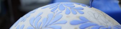 A sample image showing a craftsman adding patterns to a white jar using a brush and blue paint. The focus is on the brush, while the foreground of the jar and the background are blurred