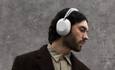 A person with a brown coat, a beard and dark hair wearing white 1000X THE COLLEXION headphones against a grey stone wall