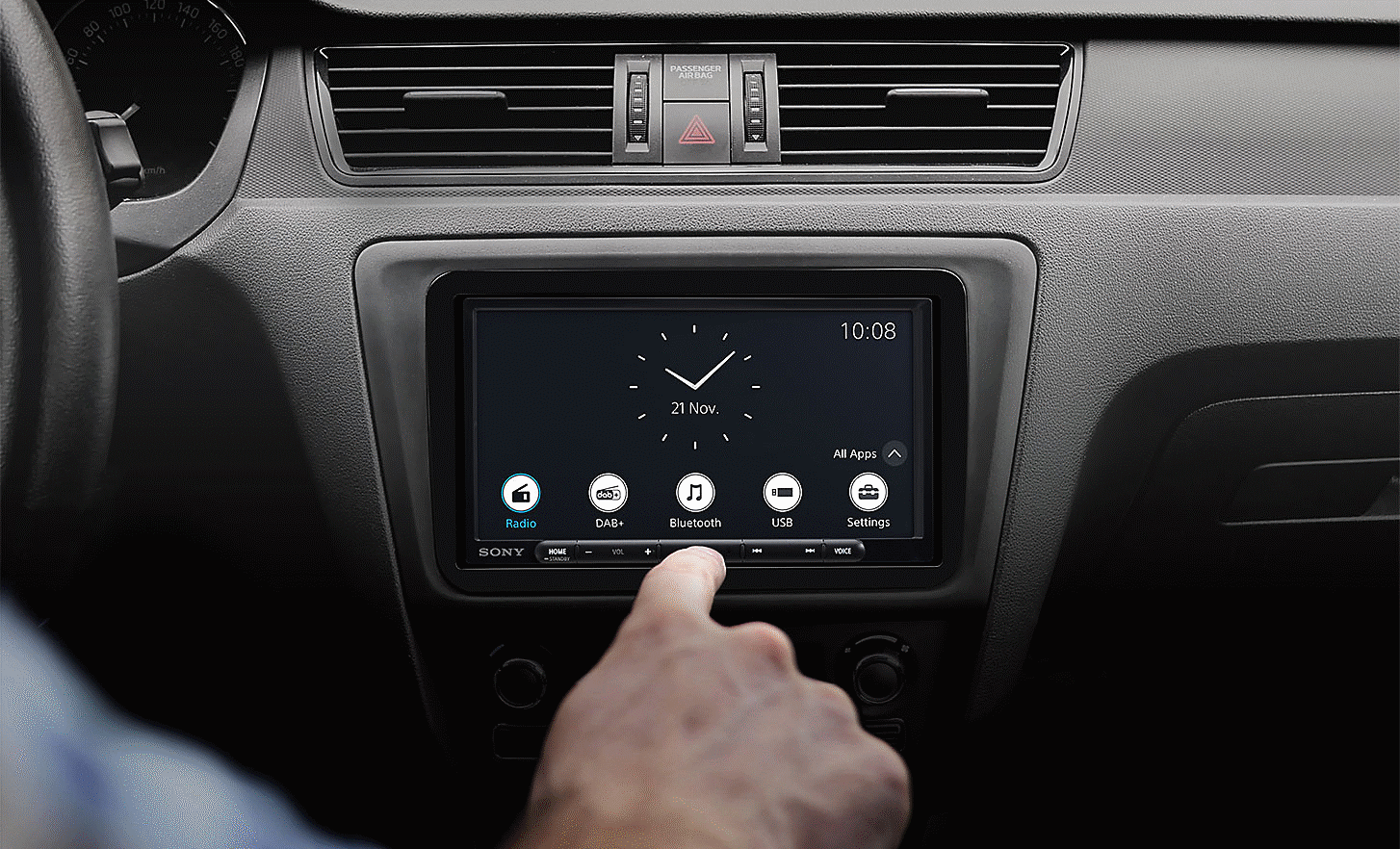 Slika uređaja XAV-AX4050 na nadzornoj ploči sa satom i više gumba na zaslonu te rukom koja lebdi u prednjem planu