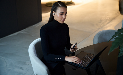 검은색 옷을 입은 젊은 여성이 화이트 이어버드를 귀에 착용하고 검은색 노트북과 휴대폰을 사용하고 있습니다.