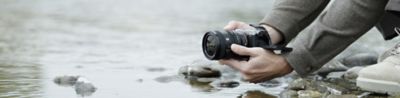 A usage image showing a user holding the α7CII with the FE 24-50mm F2.8 G near the surface of a river. The user is shooting from a low position
