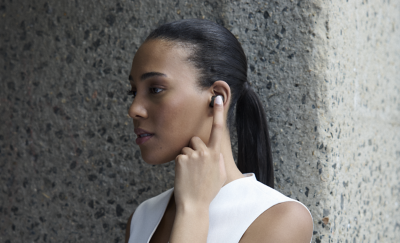 바깥에 흰색 조끼를 입은 젊은 여성이 왼쪽 귀에 블랙 이어버드를 착용하고 벽 앞에 서 있습니다. 전화를 받는 것처럼 집게손가락으로 이어버드를 터치하고 있습니다.