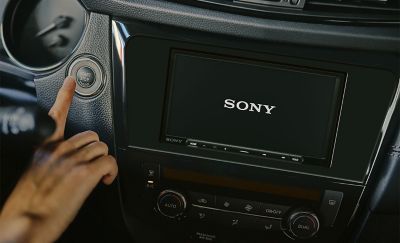 Nuotrauka – XAV-AX4050 prietaisų skydelyje ir „Sony“ logotipas ekrane, asmuo spaudžia automobilio uždegimo mygtuką.