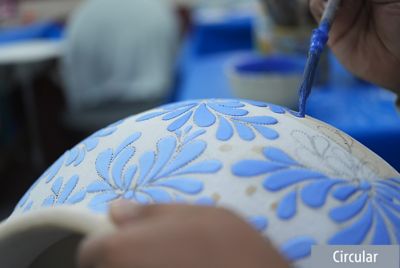 Образец изображения, на котором мастер кистью с синей краской наносит узоры на белую банку. Фокус находится на кисти, а банка и фон на заднем плане размыты