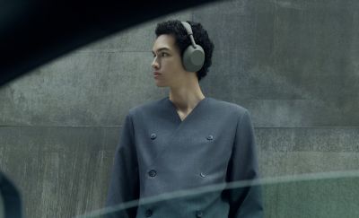 Una imagen de perfil de una persona con cabello oscuro y rizado mirando hacia la izquierda mientras usa los audífonos tipo diadema Sony WH-1000XM6 Olive Gray. Está usando una chaqueta gris de doble botonadura y está de pie frente a un muro industrial de hormigón. La imagen está tomada a través de la ventana abierta de un auto.